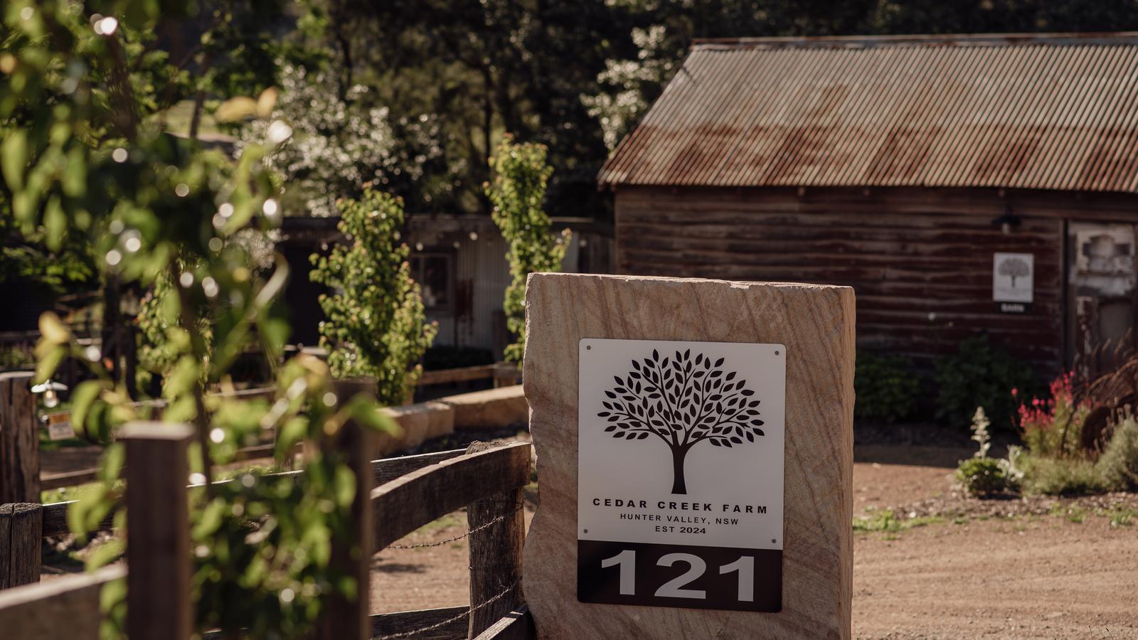 Cedar Creek Farm entry sign on a sandstone block beside a timber fence, with a corrugated-iron barn behind